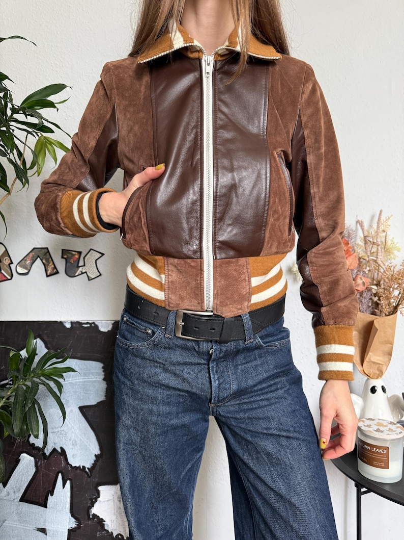 Pu&ograve; includere: Giacca bomber in pelle scamosciata e pelle marrone con cerniera bianca e polsini a righe. La giacca ha un colletto marrone e un cinturino a righe marrone e bianco. La persona indossa jeans blu scuro e una cintura nera.