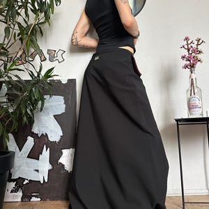 May include: A black maxi skirt with a high waist and a flowing silhouette. The skirt is paired with a black tank top. The outfit is worn with tan shoes. The background includes a mirror, artwork, and a potted plant.