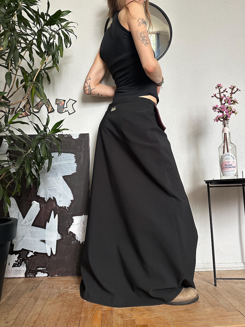 May include: A black maxi skirt with a high waist and a flowing silhouette. The skirt is paired with a black tank top. The outfit is worn with tan shoes. The background includes a mirror, artwork, and a potted plant.
