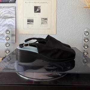 May include: Black platform sandals with a slingback strap and a light blue insole. The sandals have a buckle closure and are made of a fabric material. The shoes are on a black surface.
