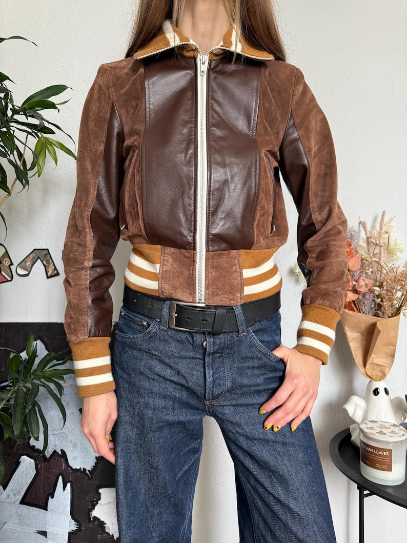 Pu&ograve; includere: Giacca in pelle marrone con zip, pannelli in camoscio e colletto e polsini a coste con strisce bianche. La giacca &egrave; abbinata a jeans blu scuro e una cintura nera.