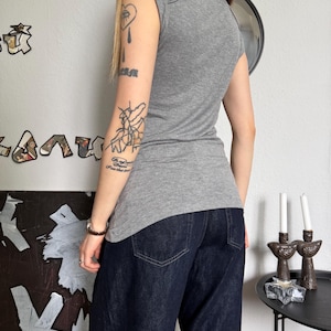 May include: A gray cap-sleeve top and dark blue denim jeans. The top has a scoop neck and a fitted silhouette. The jeans are a dark wash with a wide leg. The outfit is simple and casual.