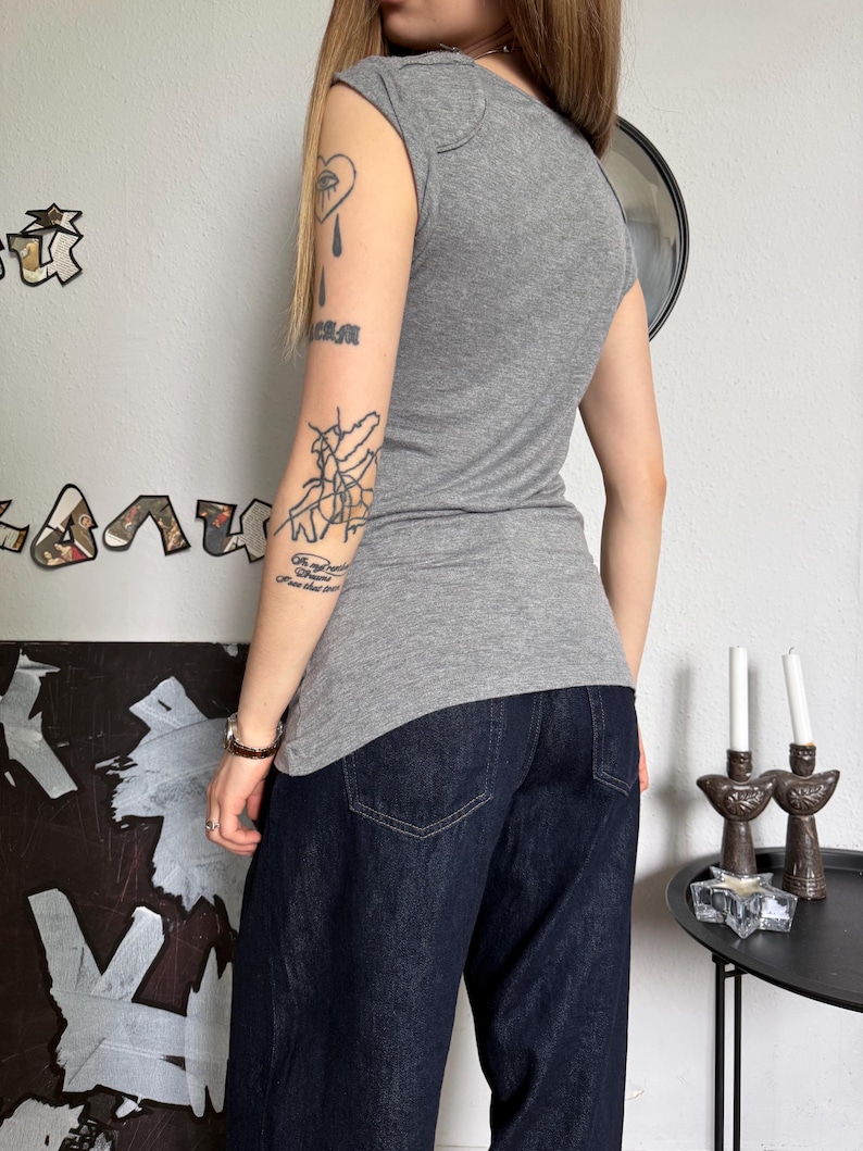 May include: A gray cap-sleeve top and dark blue denim jeans. The top has a scoop neck and a fitted silhouette. The jeans are a dark wash with a wide leg. The outfit is simple and casual.