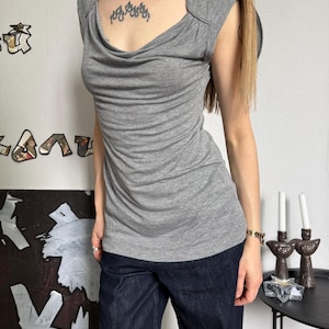 May include: A gray sleeveless top with a draped cowl neckline. The top has a fitted silhouette and is paired with dark blue denim jeans. The image shows a casual outfit suitable for everyday wear.
