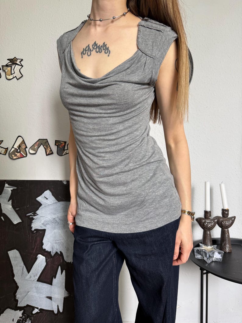 May include: A gray sleeveless top with a draped cowl neckline. The top has a fitted silhouette and is paired with dark blue denim jeans. The image shows a casual outfit suitable for everyday wear.