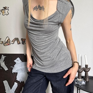 May include: A gray, short-sleeved top with a draped neckline and a fitted silhouette. The top is paired with dark blue denim pants. The top has a cowl neck and shoulder detailing. The overall style is casual and chic.