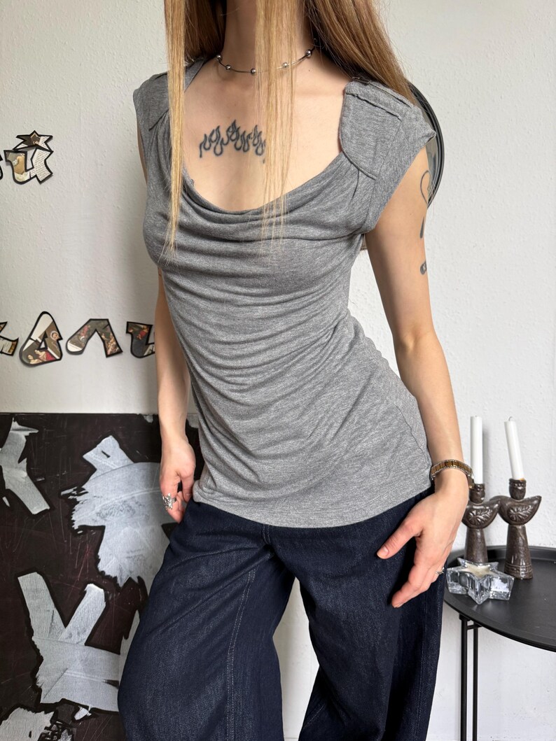 May include: A gray, short-sleeved top with a draped neckline and a fitted silhouette. The top is paired with dark blue denim pants. The top has a cowl neck and shoulder detailing. The overall style is casual and chic.