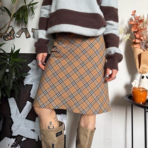May include: A tan plaid skirt with a brown, black, and white pattern. The skirt is paired with a striped brown, grey, and light blue jumper. The outfit is completed with tan boots.