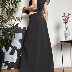 May include: A black maxi skirt with a high waist and a slight A-line silhouette. The skirt features decorative silver-toned buttons and a small pocket detail. The model is wearing a black tank top. The skirt is made of a smooth, opaque fabric.