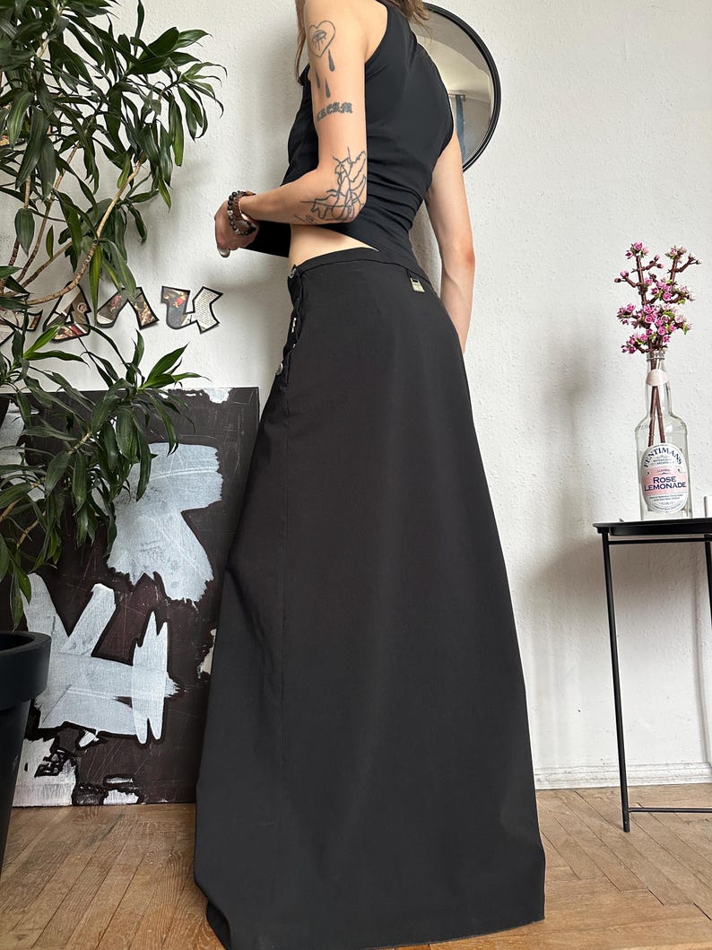 May include: A black maxi skirt with a high waist and a slight A-line silhouette. The skirt features decorative silver-toned buttons and a small pocket detail. The model is wearing a black tank top. The skirt is made of a smooth, opaque fabric.
