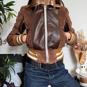 Pu&ograve; includere: Giacca in pelle scamosciata e pelle marrone con zip. La giacca presenta un pannello centrale in pelle marrone e maniche in pelle scamosciata marrone. I polsini e la vita hanno un design a maglia a coste con strisce bianche e marroni. La giacca ha due tasche anteriori.