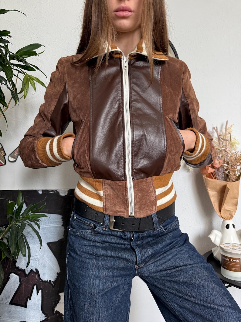 Pu&ograve; includere: Giacca in pelle scamosciata e pelle marrone con zip. La giacca presenta un pannello centrale in pelle marrone e maniche in pelle scamosciata marrone. I polsini e la vita hanno un design a maglia a coste con strisce bianche e marroni. La giacca ha due tasche anteriori.