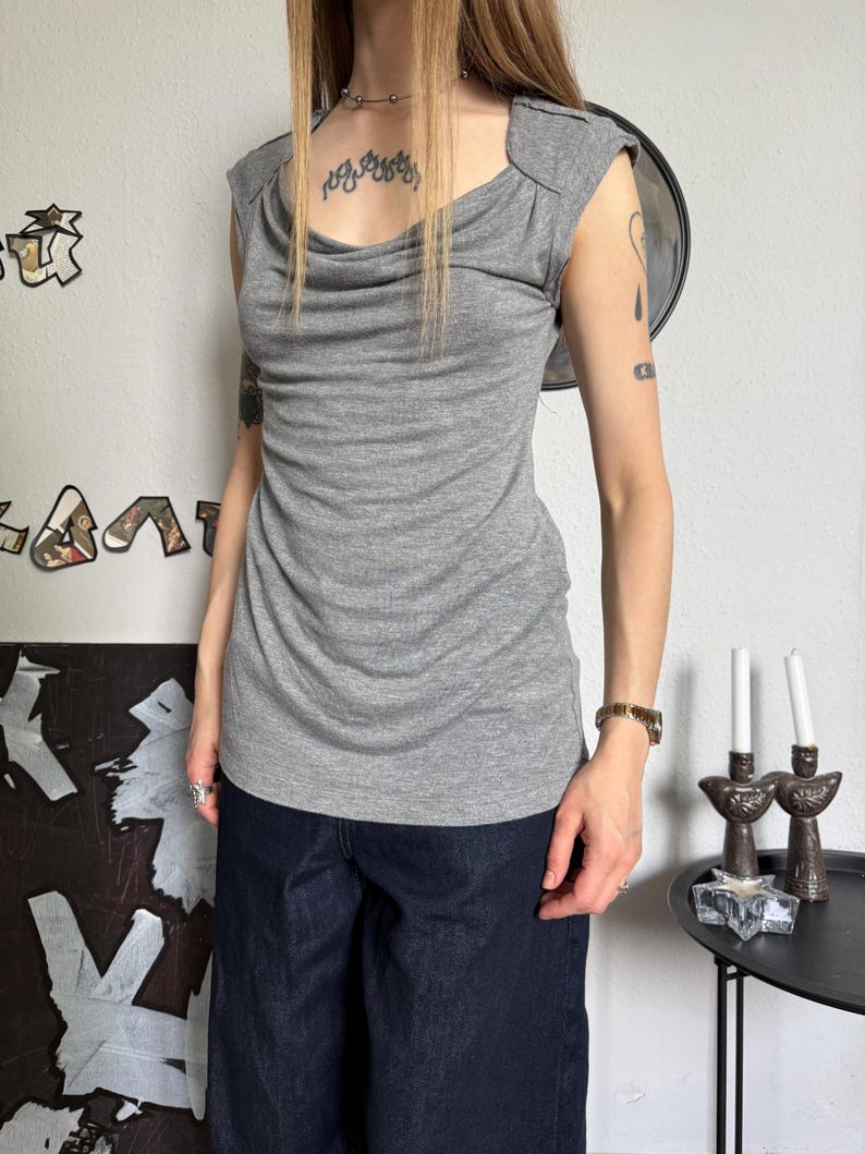 May include: A gray, sleeveless top with a draped cowl neckline. The top has a fitted silhouette and shoulder detailing. The model is wearing dark blue denim pants. The background includes wall art and a small side table with candles.