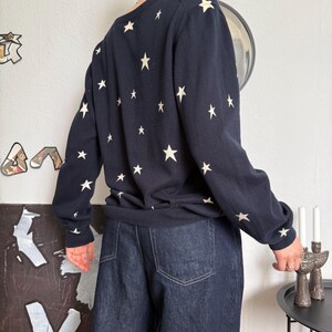 以下が含まれることがあります： 背中と袖に小さなオフホワイトの星が繰り返しプリントされた長袖のネイビーブルーのセーターと、後ろポケットにステッチが施されたダークブルーのデニムジーンズの組み合わせ。