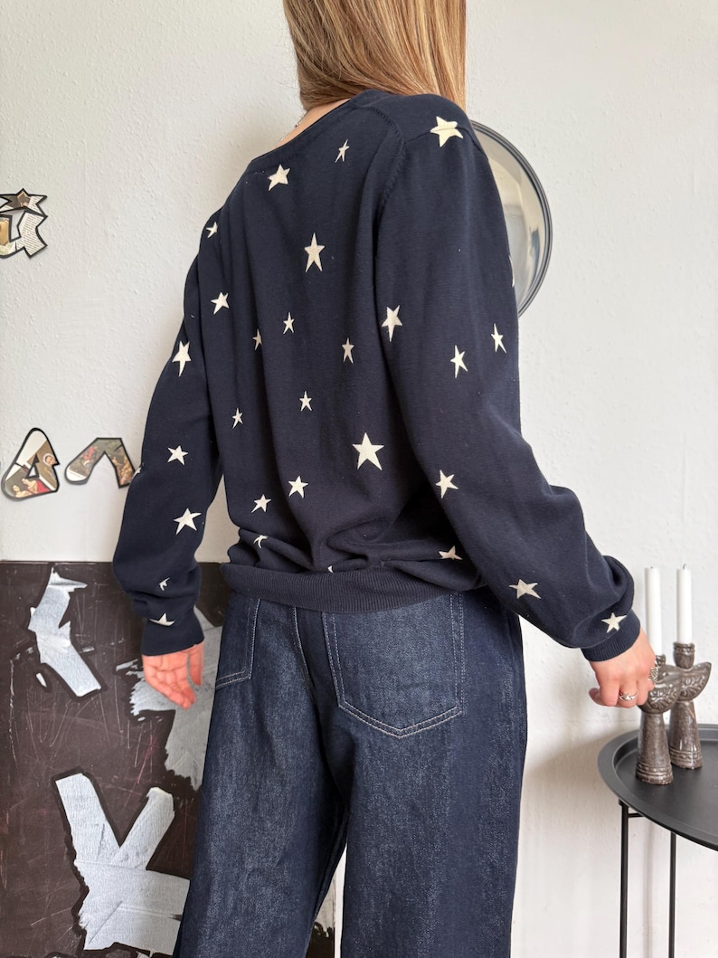 以下が含まれることがあります： 背中と袖に小さなオフホワイトの星が繰り返しプリントされた長袖のネイビーブルーのセーターと、後ろポケットにステッチが施されたダークブルーのデニムジーンズの組み合わせ。