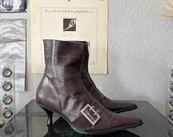 May include: A pair of brown leather ankle boots with pointed toes and a small heel. The boots feature a silver buckle detail on the side and a zipper closure. The boots are displayed on a reflective surface.