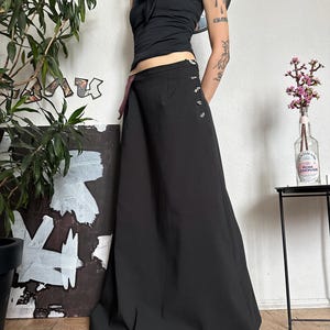 May include: A black halter top with a bow detail and a long black skirt. The skirt has decorative buttons on the side. The outfit is worn with a black choker. A plant and artwork are in the background.