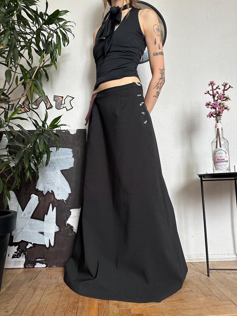 May include: A black halter top with a bow detail and a long black skirt. The skirt has decorative buttons on the side. The outfit is worn with a black choker. A plant and artwork are in the background.