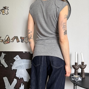 May include: A gray cap-sleeve top and dark blue denim jeans. The top has a scoop neck and shoulder detailing. The jeans are a wide-leg style. The person is standing in front of a white wall with art and a mirror.