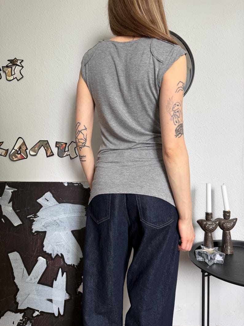 May include: A gray cap-sleeve top and dark blue denim jeans. The top has a scoop neck and shoulder detailing. The jeans are a wide-leg style. The person is standing in front of a white wall with art and a mirror.