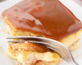 Op de afbeelding: Een vierkant gebakje met lagen bladerdeeg en een romige vulling, gegarneerd met een glanzende karamelsaus. Een vork wordt in het dessert gestoken, waardoor de lagen zichtbaar worden. Het dessert ligt op een wit bord.