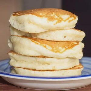 Puede incluir: Una pila de cinco panqueques esponjosos en un plato blanco con un borde azul. Los panqueques son de color marrón dorado en la parte superior y de color claro. El plato está sobre una superficie de madera.