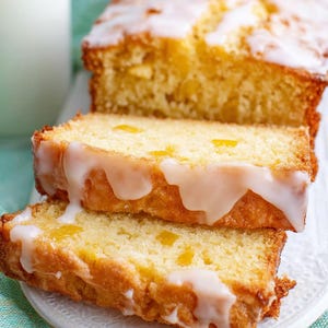 Op de afbeelding: Een plakje citroenbrood met witte glazuur, gepresenteerd op een wit bord. Het brood heeft een goudgele kleur en zichtbare stukjes citroen. Een glas melk is zichtbaar op de achtergrond.