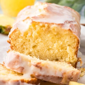 May include: A loaf of yellow cake with a white glaze, partially sliced to reveal the moist interior. The cake is on a white surface, with a blurred background including a pineapple and green leaves.