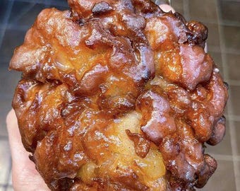 May include: A close-up of a large, golden-brown fritter, with a glossy, glazed surface. The fritter has an irregular shape with many peaks and valleys, suggesting a crispy exterior and a soft interior. The colors range from light tan to deep brown.