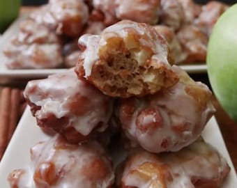 May include: A stack of apple fritters on a white plate. The fritters are golden brown with a white glaze. One fritter is partially bitten, revealing the apple filling. Green apples are visible in the background.