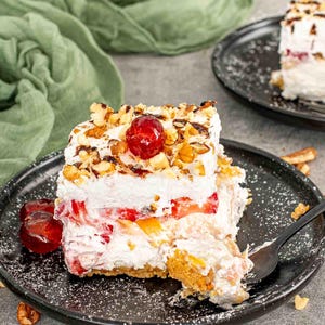 Puede incluir: Una porción de postre en capas sobre un plato negro. El postre presenta una base de galleta, una capa de fruta y crema batida, cubierta con nueces, salsa de chocolate y una cereza. Hay un tenedor en el postre.