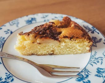 May include: A slice of apple cake with a golden-brown crust sits on a white plate with a blue floral design. A silver fork rests on the plate, ready to enjoy the dessert. The cake has a light yellow interior and a dark brown topping.