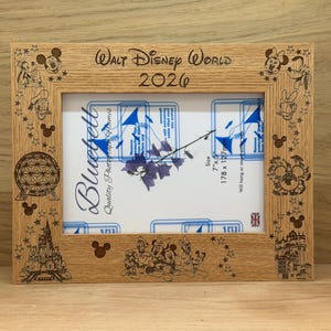 Op de afbeelding: Een rechthoekige fotolijst van lichtbruin hout met in laser gegraveerde Disney-figuren en bezienswaardigheden. De bovenrand toont de tekst Walt Disney World 2026. De lijst bevat een witte inleg met blauwe tekst en een bloemafbeelding.