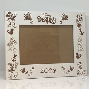 Può includere: Una cornice bianca con illustrazioni marroni di personaggi Disney, tra cui Topolino e Minnie. La cornice presenta la scritta "Disney Destiny" e l'anno "2026". La cornice è di forma rettangolare.