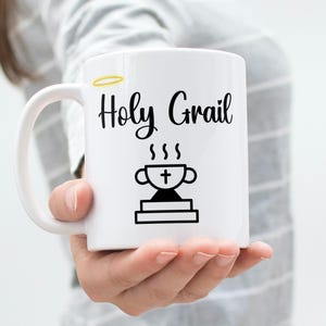May include: White ceramic mug with a black design that says "Holy Grail" with a stylized image of a chalice with a cross on top and steam coming from it.