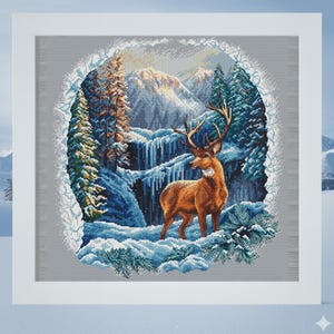 May include: A framed cross-stitch depicting a winter landscape. A brown deer stands in the snow, with a waterfall and snow-covered evergreen trees in the background. The scene is framed by a white border and set against a light blue backdrop.