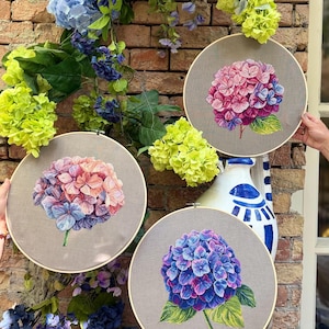 Puede incluir: Tres bastidores de bordado circulares de madera con paneles de tela, cada uno con una flor de hortensia bordada a mano. El bastidor superior derecho muestra una flor rosa y morada, el izquierdo una flor color melocotón y azul, y el inferior una flor azul oscuro.