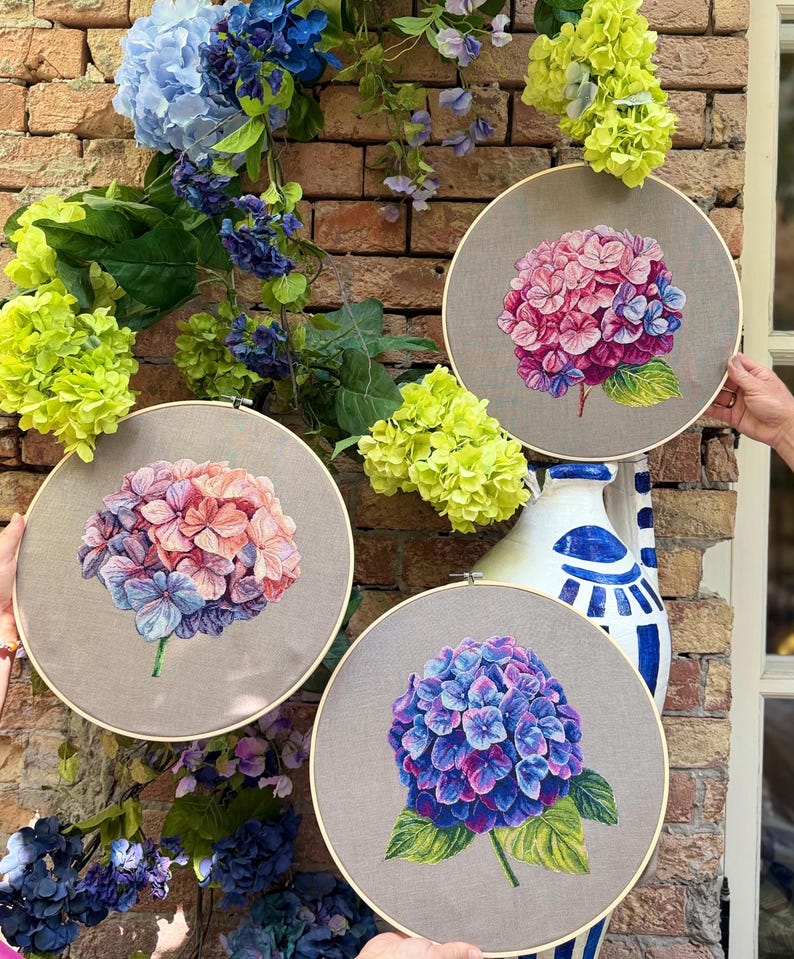 Puede incluir: Tres bastidores de bordado circulares de madera con paneles de tela, cada uno con una flor de hortensia bordada a mano. El bastidor superior derecho muestra una flor rosa y morada, el izquierdo una flor color melocot&oacute;n y azul, y el inferior una flor azul oscuro.