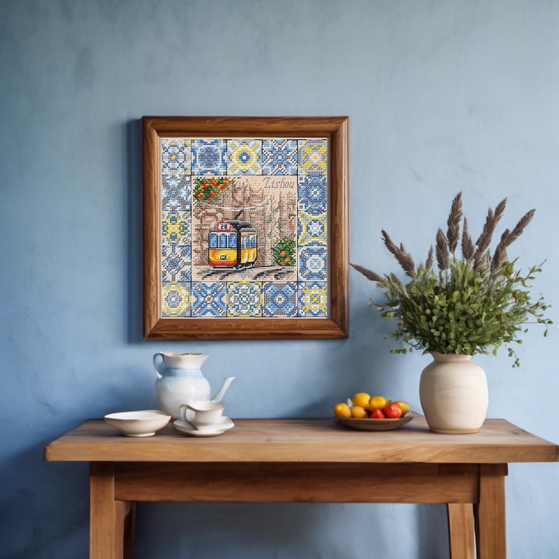 May include: A framed cross-stitch depicting a yellow and red tram in Lisbon, Portugal, with a stone wall background and blue and yellow tile patterns. The wooden frame is on a wooden table with a vase of greenery and a bowl of fruit.