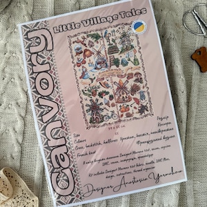 Op de afbeelding: Een kruissteekpakket getiteld "Little Village Tales" met een gedetailleerde dorpsscène met windmolens, pompoenen en andere herfstachtige elementen. De kit bevat Zweigart Murano 32ct stof, een naald, DMC-garen en instructies. De afmetingen zijn 39 x 55 cm.