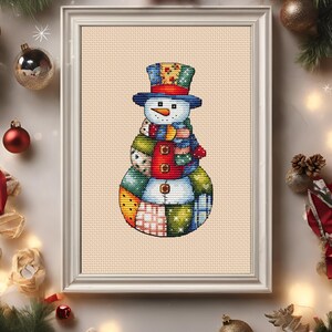 Pode incluir: Arte em ponto cruz emoldurada de um boneco de neve. O boneco de neve tem um design de patchwork com um chap&eacute;u alto azul, um cachecol azul e um colete vermelho. O fundo &eacute; bege claro.