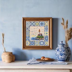 May include: A framed cross-stitch picture depicting a lighthouse on a rocky outcrop, surrounded by a blue and yellow tile design. The lighthouse has a red top and is set against a blue sky with birds. The frame is wooden.