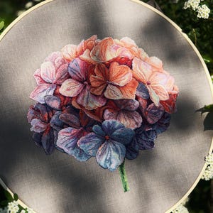 Puede incluir: Un bastidor de bordado circular de madera que contiene un dise&ntilde;o de punto de cruz de una flor de hortensia. La flor presenta p&eacute;talos en tonos rosa, naranja, morado y azul, sobre un fondo de tela gris s&oacute;lido con un solo tallo verde.