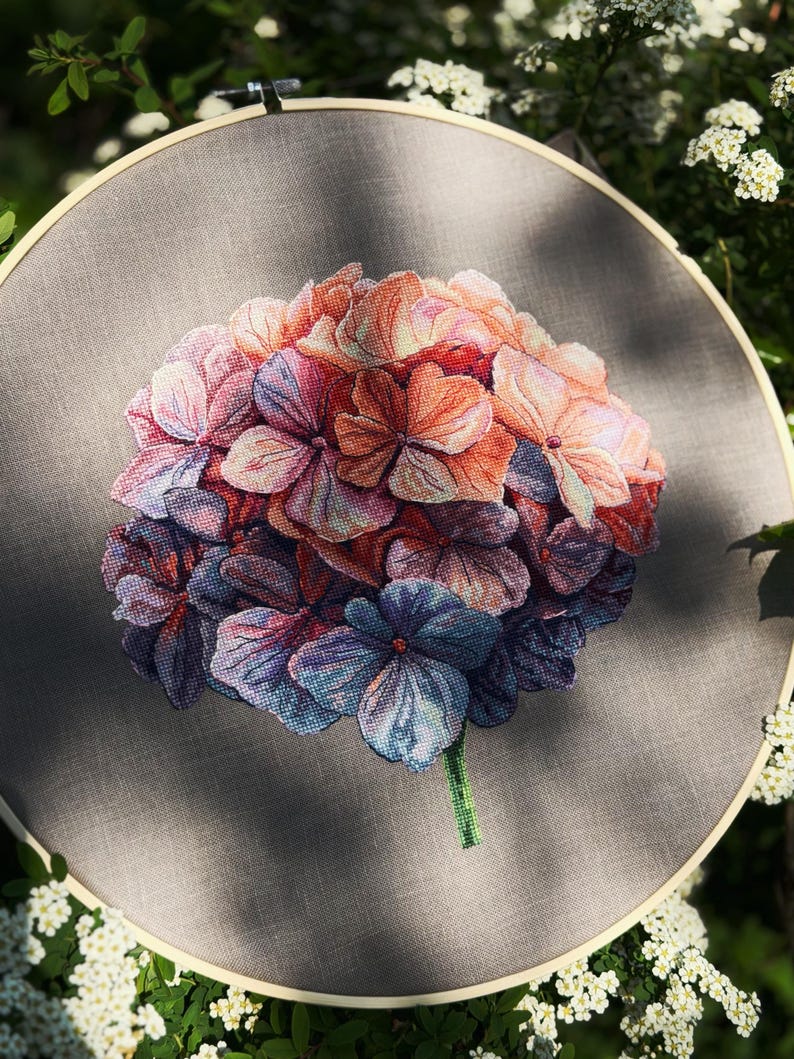 Puede incluir: Un bastidor de bordado circular de madera que contiene un dise&ntilde;o de punto de cruz de una flor de hortensia. La flor presenta p&eacute;talos en tonos rosa, naranja, morado y azul, sobre un fondo de tela gris s&oacute;lido con un solo tallo verde.