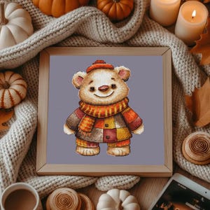 May include: A framed cross-stitch of a smiling teddy bear wearing a hat, scarf, and patchwork coat in shades of brown, orange, and yellow. The bear is centered on a light purple background, surrounded by a wooden frame.