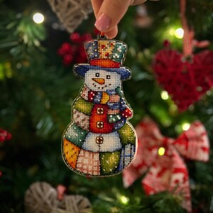 Pode incluir: Enfeite de Natal de boneco de neve bordado em ponto cruz. O boneco de neve tem um chap&eacute;u azul, um nariz de cenoura e um cachecol e corpo de retalhos. O enfeite &eacute; segurado em frente a uma &aacute;rvore de Natal.