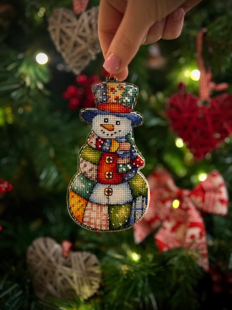 Pode incluir: Enfeite de Natal de boneco de neve bordado em ponto cruz. O boneco de neve tem um chap&eacute;u azul, um nariz de cenoura e um cachecol e corpo de retalhos. O enfeite &eacute; segurado em frente a uma &aacute;rvore de Natal.