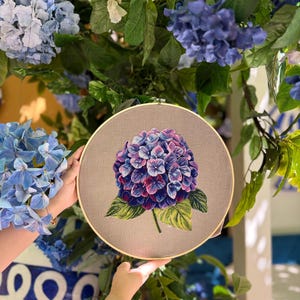 Puede incluir: Un bastidor de bordado circular de madera que contiene una tela de color marr&oacute;n claro. Centrada en la tela hay una flor de hortensia bordada con hilo, detallada con p&eacute;talos en tonos vibrantes de morado, azul y rosa y hojas verdes.