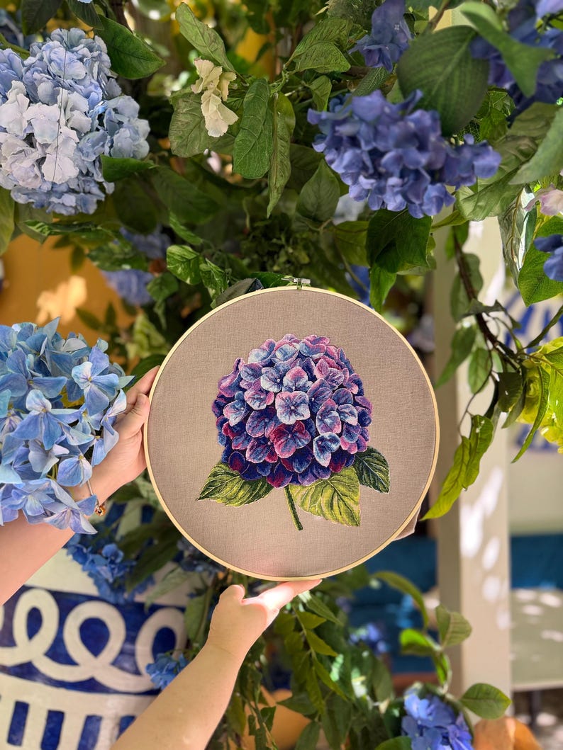 Puede incluir: Un bastidor de bordado circular de madera que contiene una tela de color marr&oacute;n claro. Centrada en la tela hay una flor de hortensia bordada con hilo, detallada con p&eacute;talos en tonos vibrantes de morado, azul y rosa y hojas verdes.