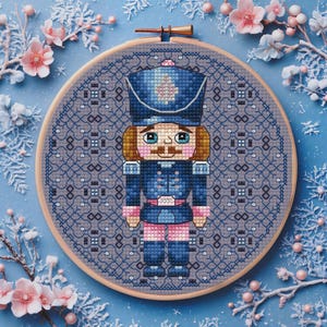 May include: A cross-stitch embroidery of a nutcracker soldier in a blue uniform, pink accents, and a brown mustache. The embroidery is set in a wooden hoop against a light blue background with floral and snowflake decorations.